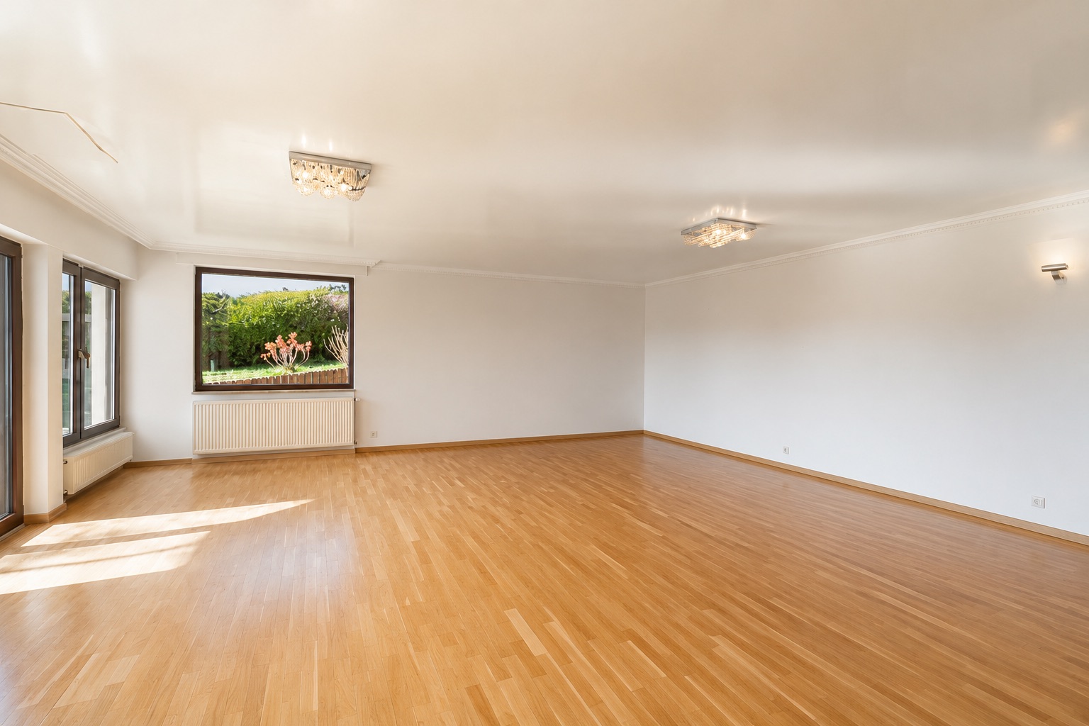 Großes, leeres Wohnzimmer mit Parkettboden, weißen Wänden und großen Fenstern mit Blick ins Grüne.