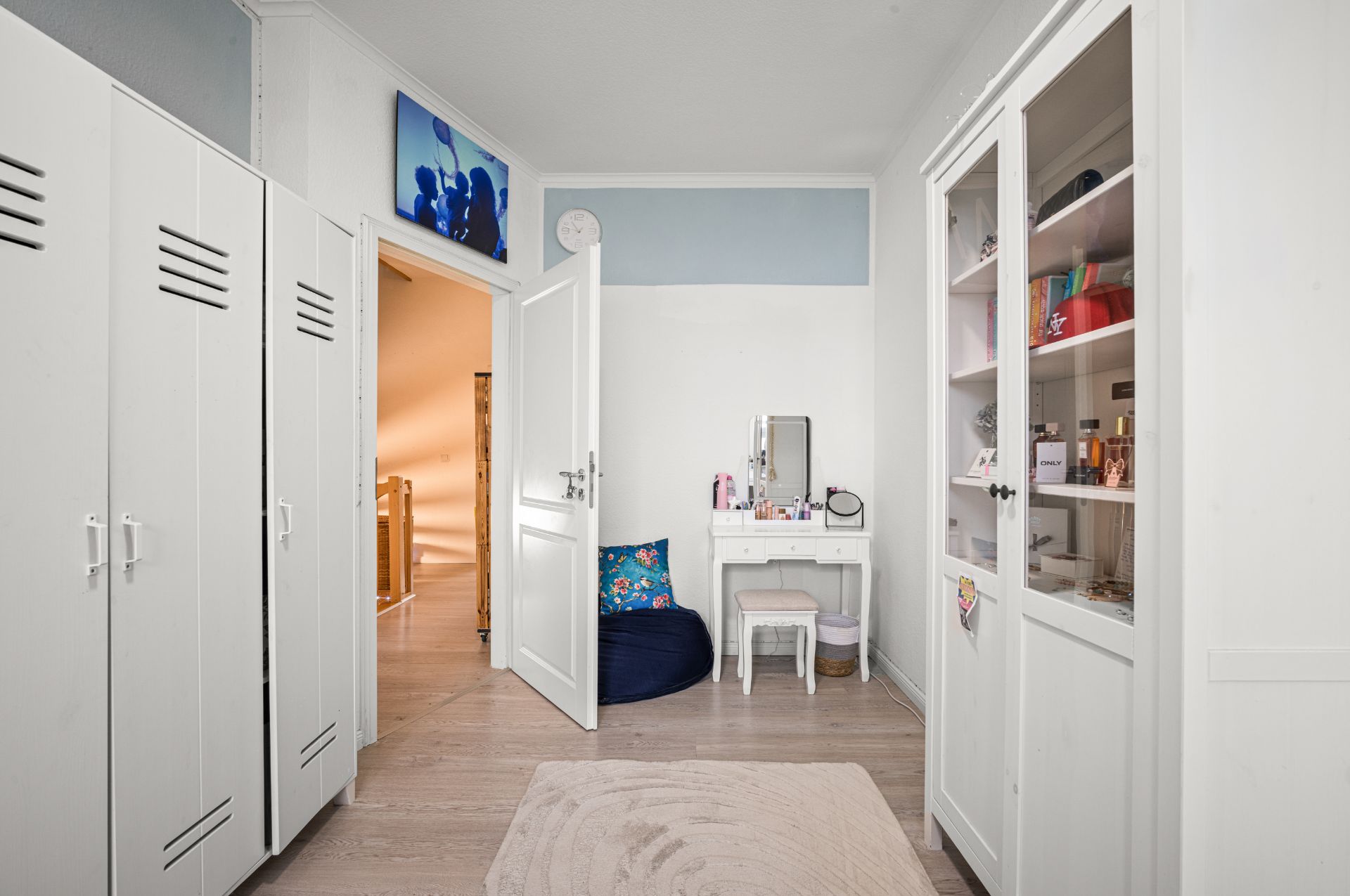 Helles Zimmer mit weißen Schränken, einem Schminktisch, Regal und offen stehender Tür.