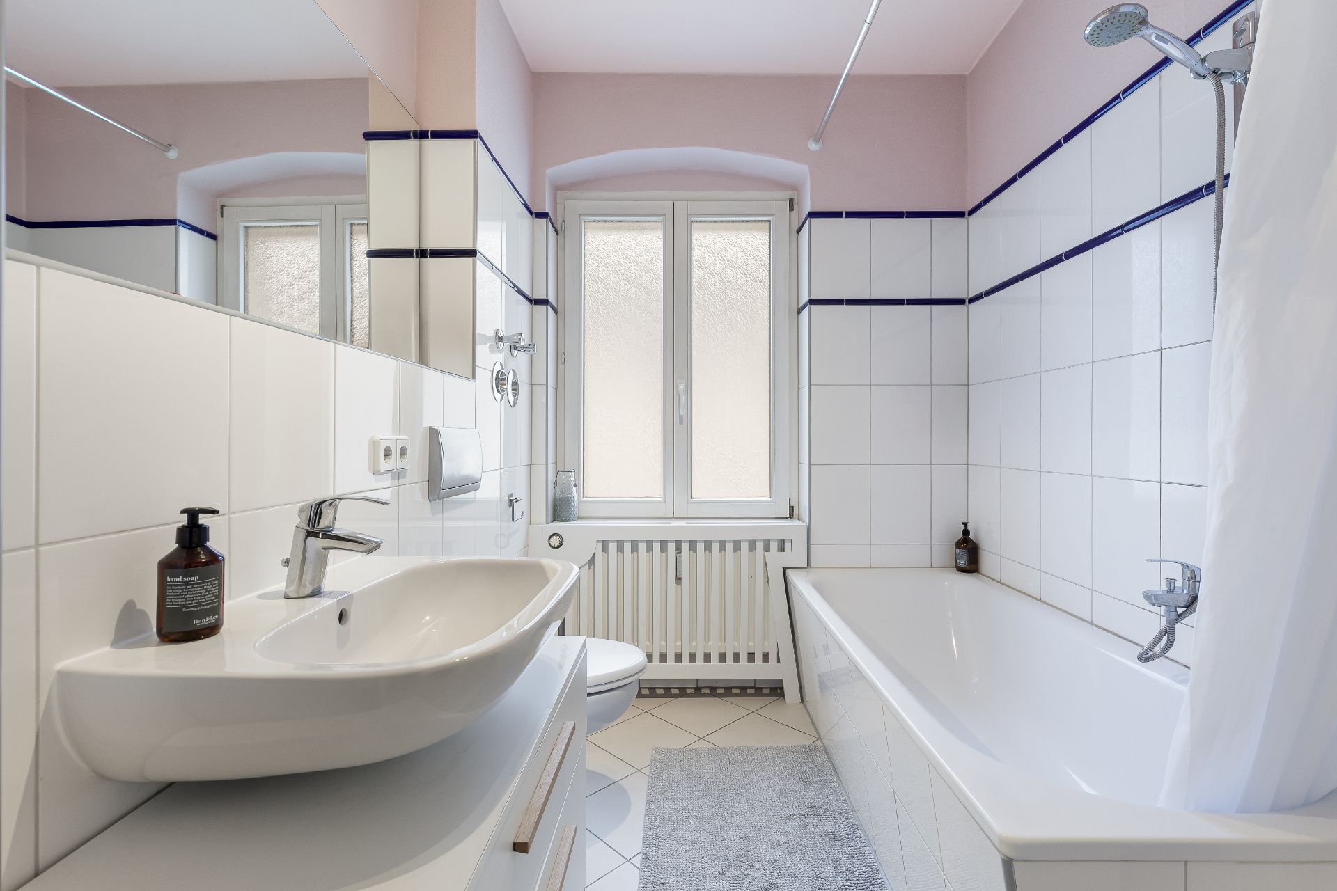 Helles Badezimmer mit Waschbecken, Badewanne, Dusche, Fenster und Fliesenboden.