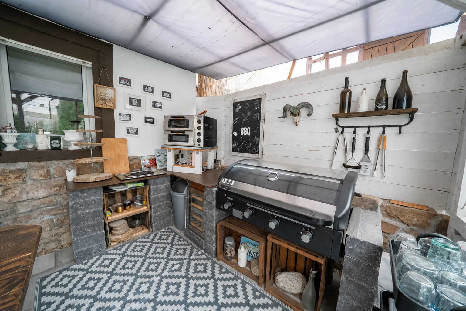 Überdachte Außenküche mit Grill, Regalen, Küchenutensilien und gemustertem Teppich.