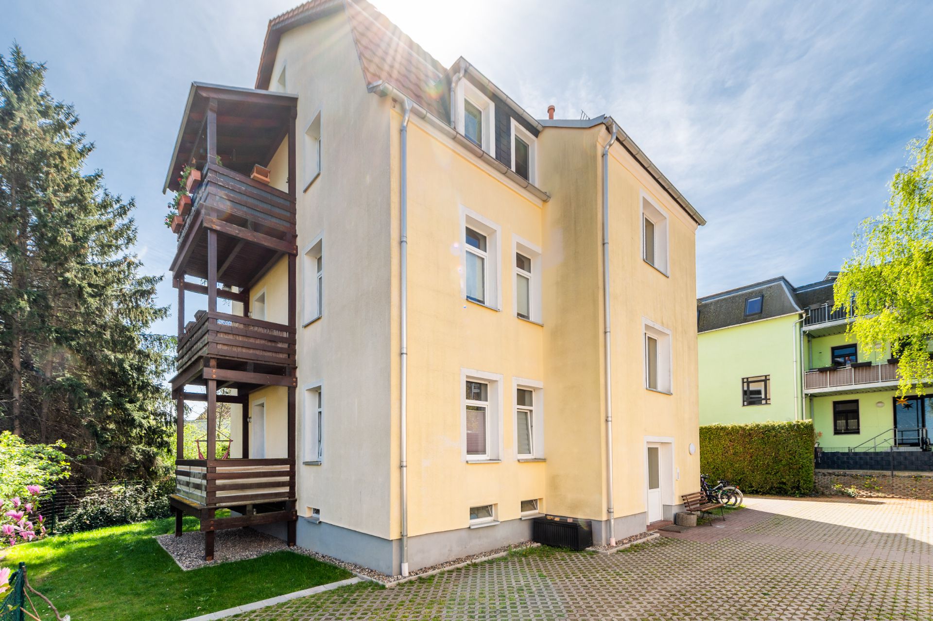 Gelbes Mehrfamilienhaus mit Holz-Balkonen und gepflastertem Hof im Grünen.