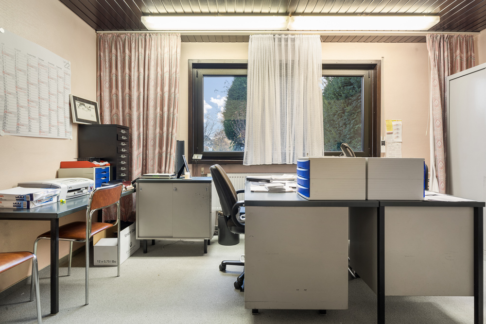 Büro mit Schreibtischen, Stühlen, Regalen, Computer und Fenster mit Vorhängen.