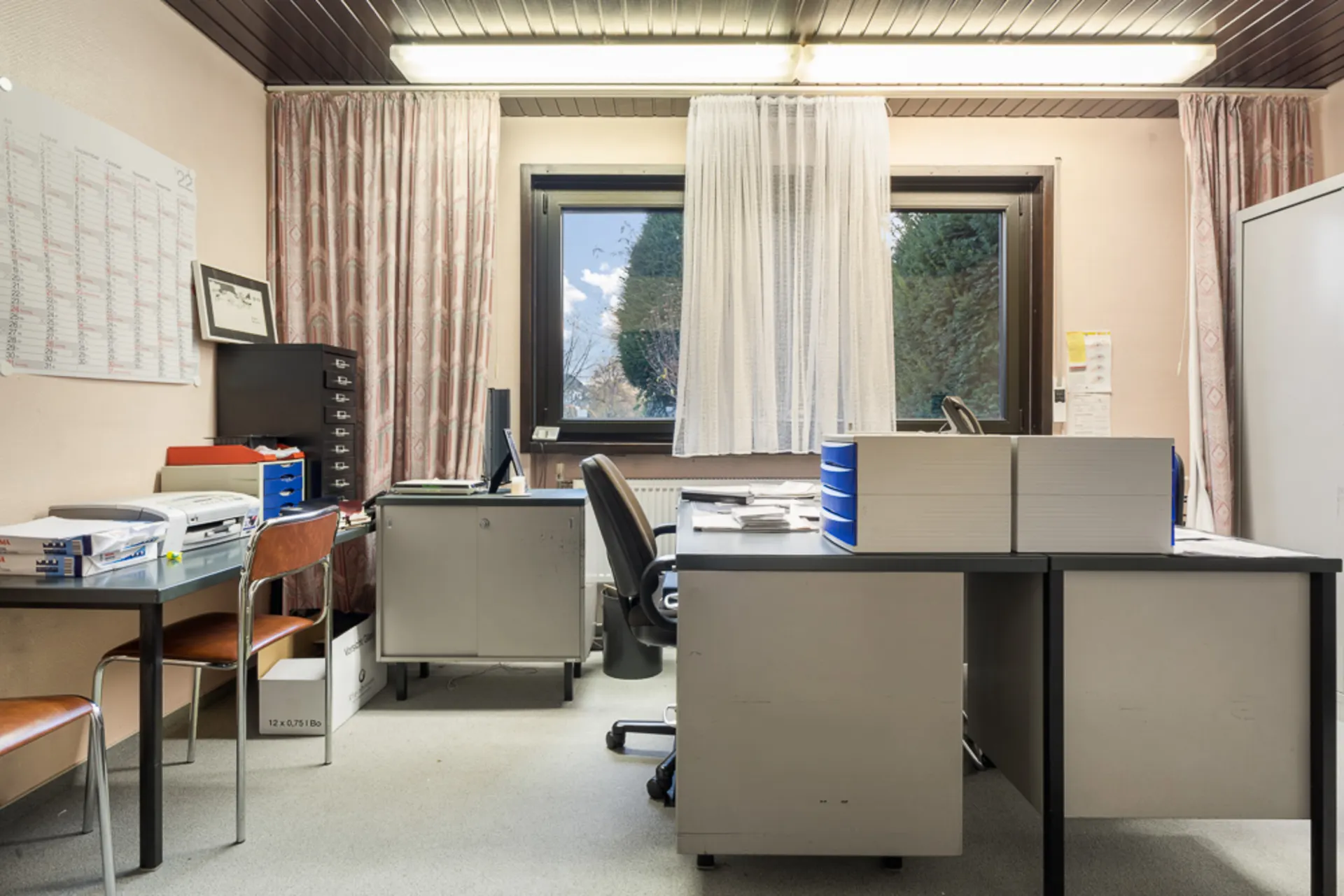 Büro mit Schreibtischen, Stühlen, Regalen, Computer und Fenster mit Vorhängen.