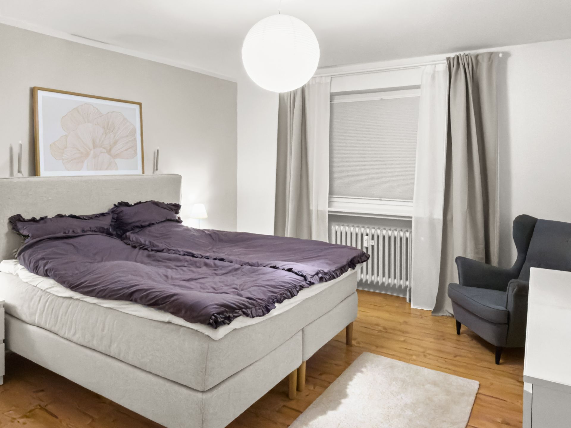 Modernes Schlafzimmer mit Doppelbett, Sessel, Fenster mit Vorhängen und Holzfußboden.
