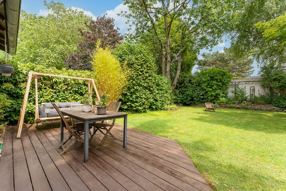Garten mit Holzterrasse, Tisch, Stühlen und einer Holzschaukelbank, umgeben von Bäumen und Sträuchern.