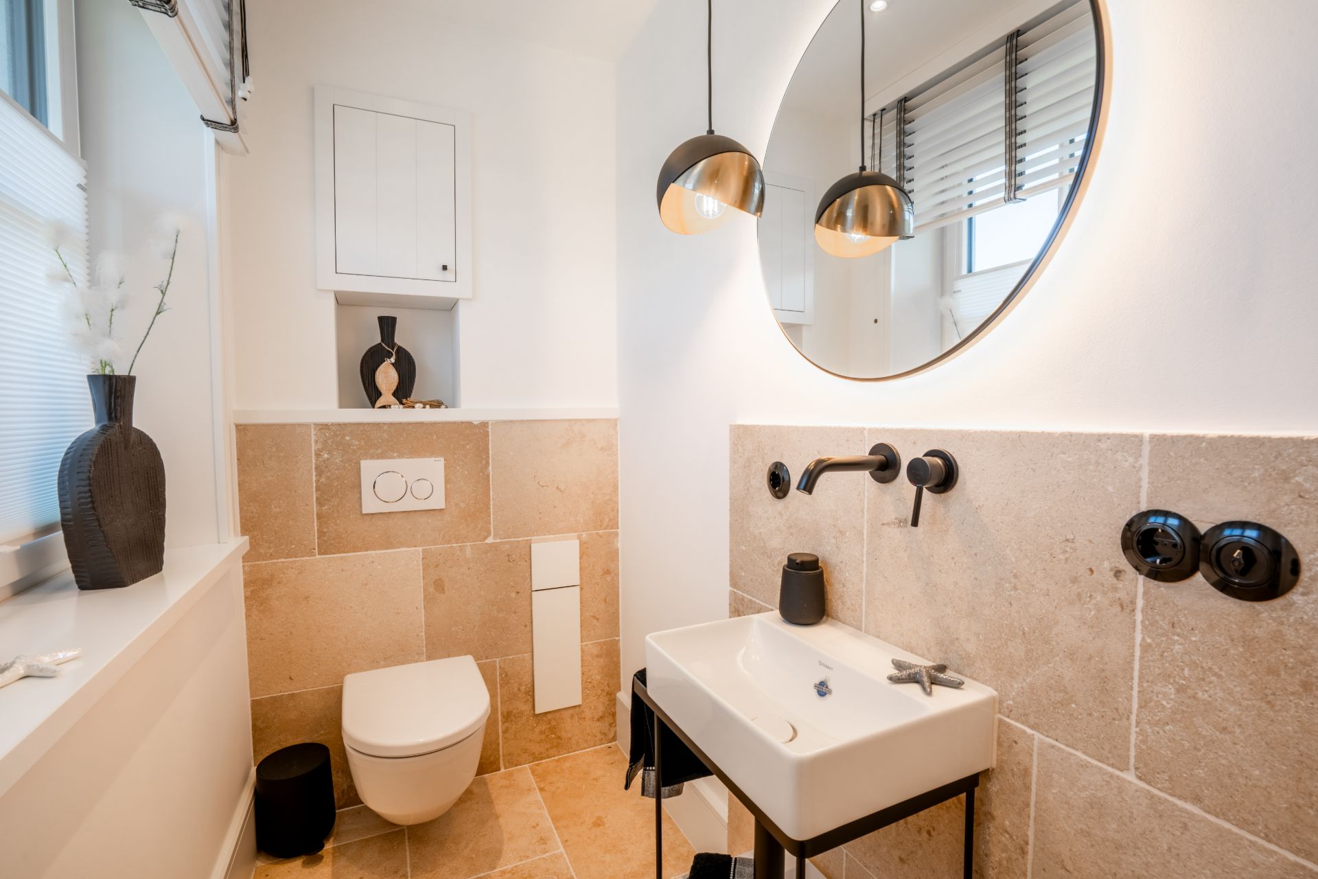 Modernes Badezimmer mit Wandtoilette, Waschbecken, rundem Spiegel und zwei Hängelampen.