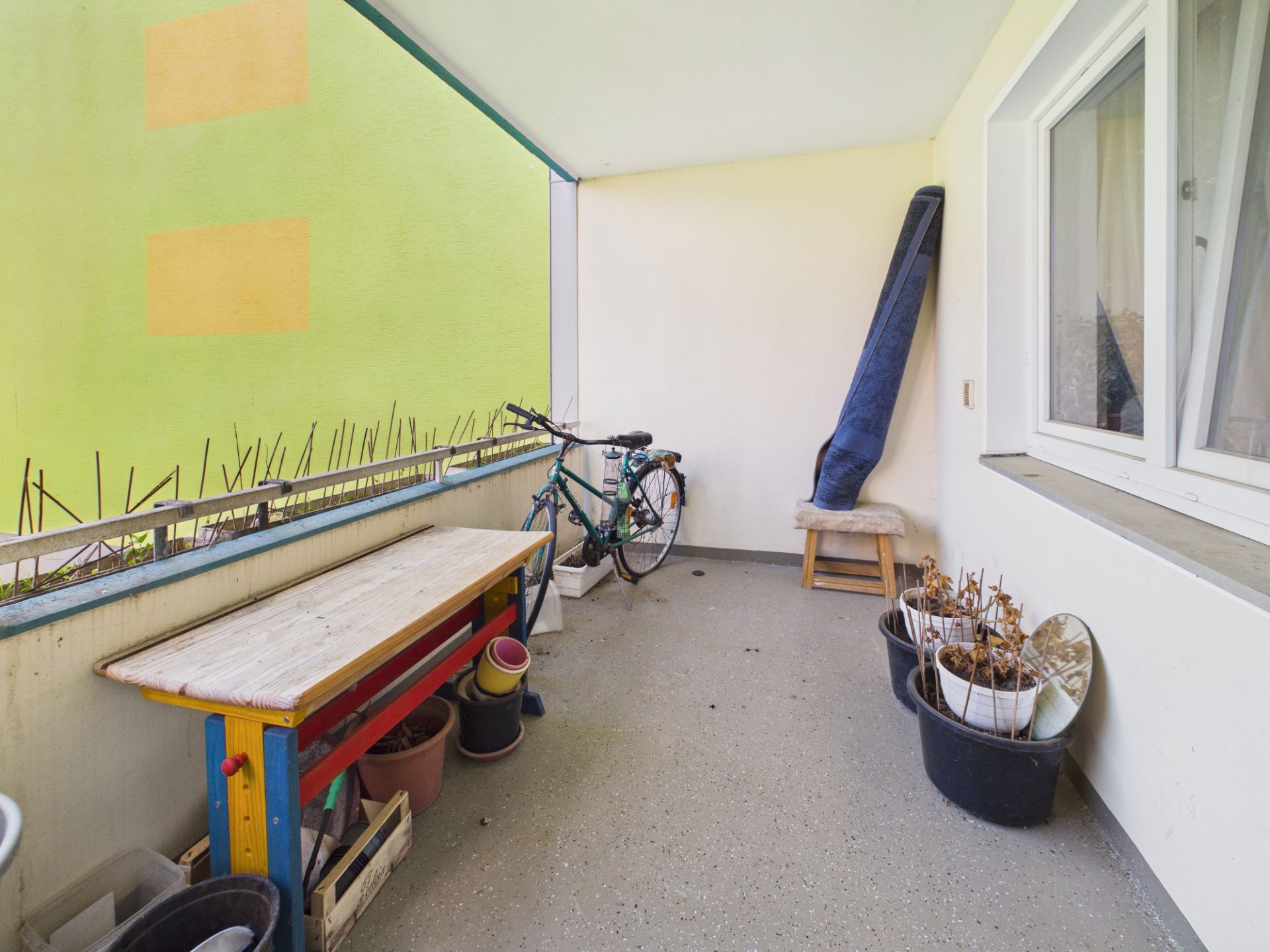 Überdachter Balkon mit Tisch, Blumentöpfen, Fahrrad und Hocker.