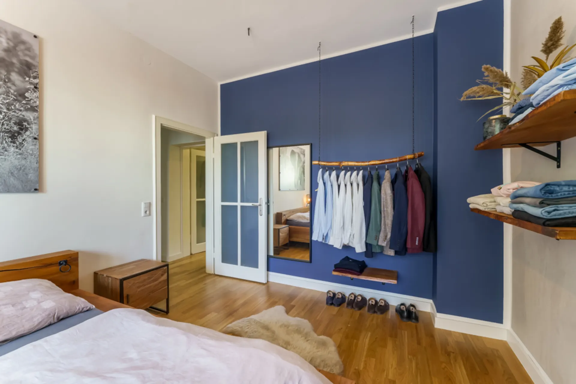Schlafzimmer mit Holzboden, Bett, blauer Wand, Kleiderstange, Spiegel und Regal.