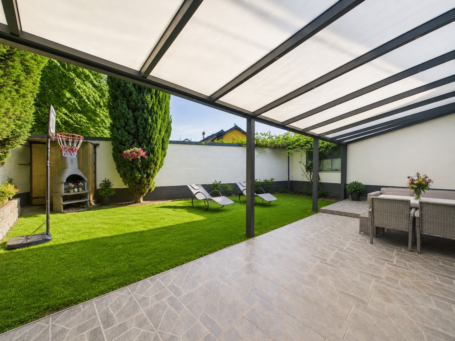 Überdachte Terrasse mit Sitzbereich, Blick auf Garten mit Liegestühlen und Basketballkorb.
