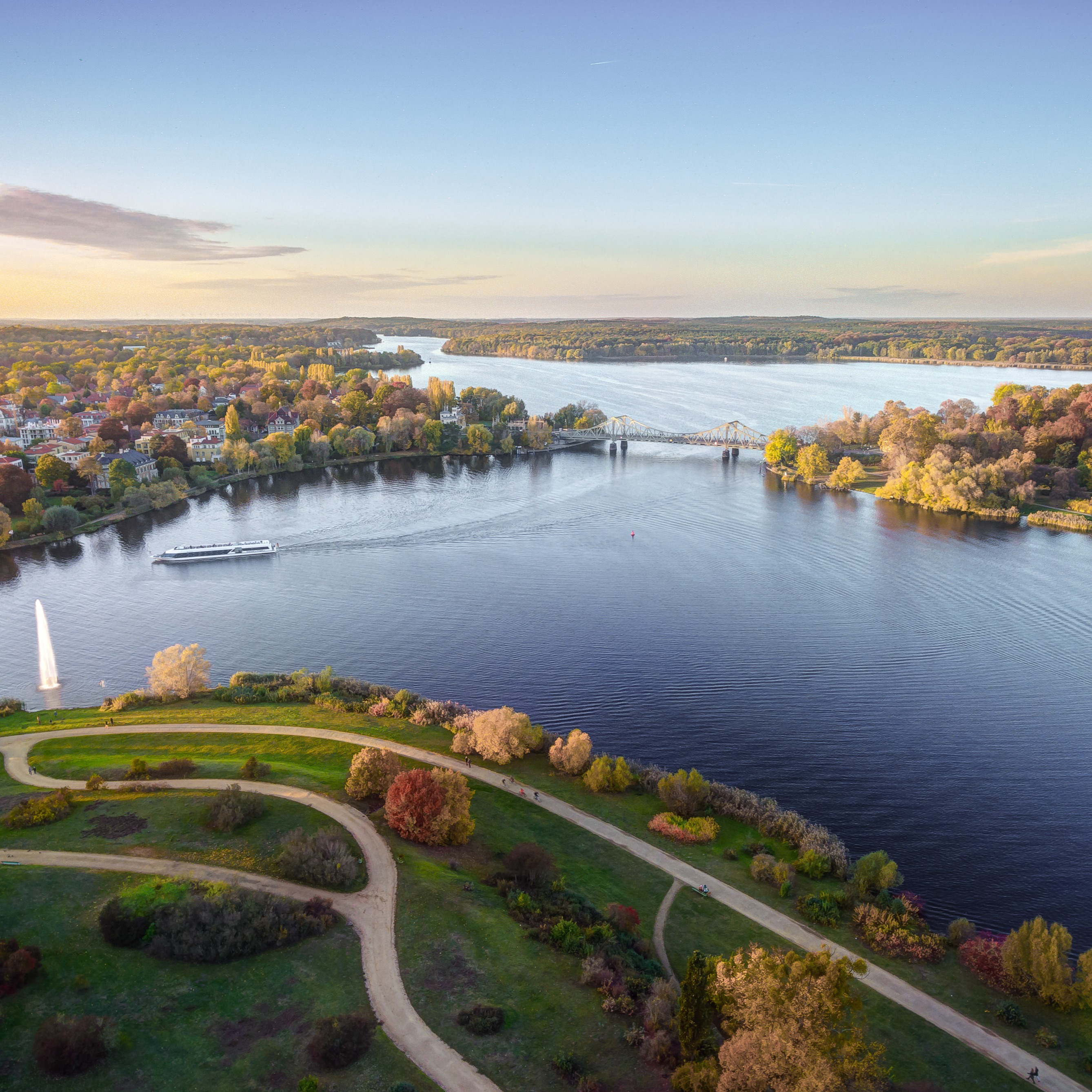 Luftaufnahme eines Parks mit Wegen am Wasser, einer Fontäne, Bäumen und einer Brücke im Hintergrund.