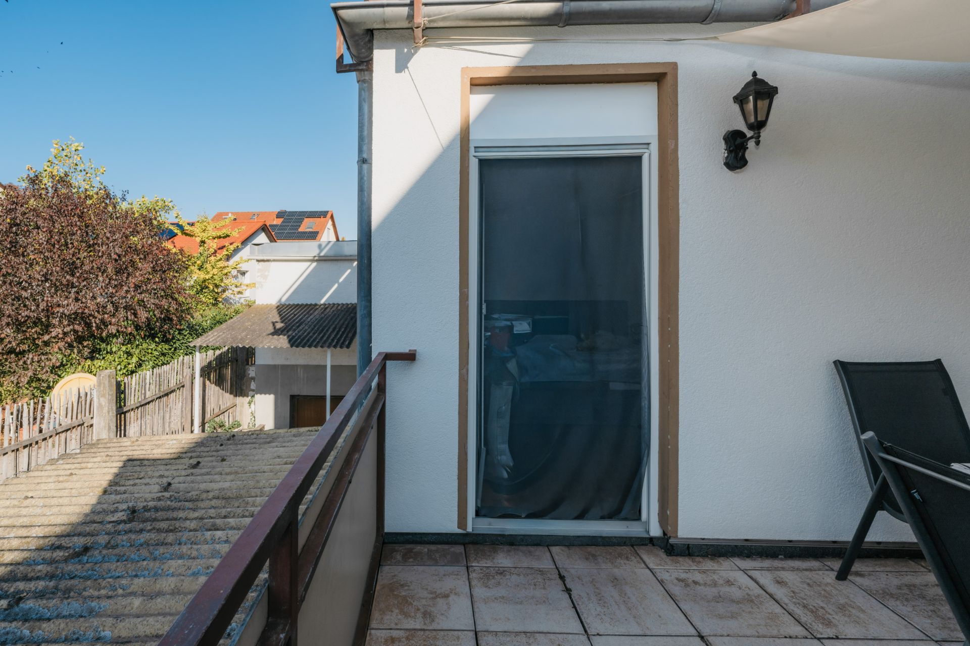 Balkon mit Fliesenboden, Stuhl, Tür und Außenleuchte an weißer Wand.