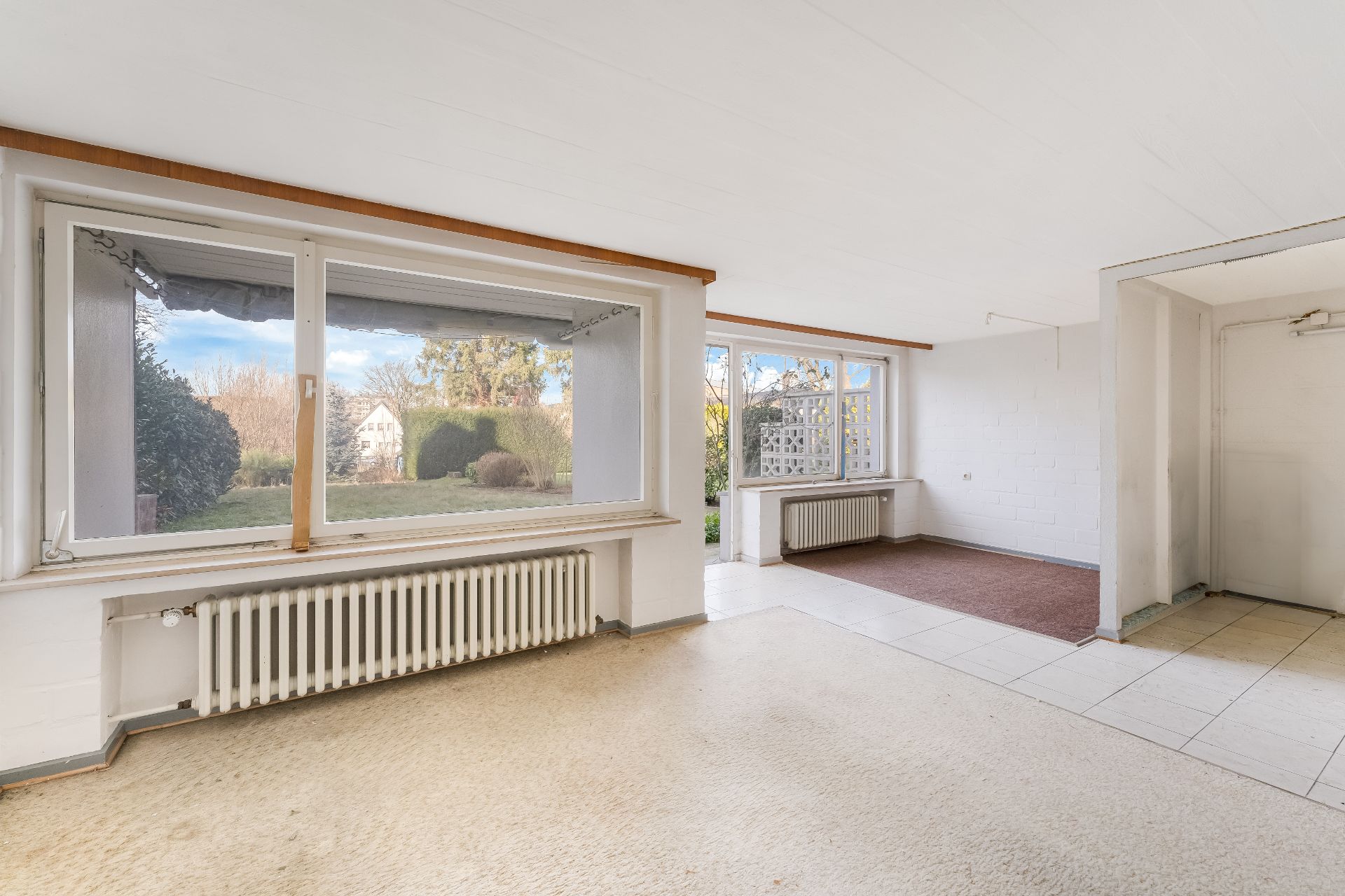 Heller Raum mit großen Fenstern, Blick auf Garten, Heizkörper unter den Fenstern, gefliester und Teppichboden.
