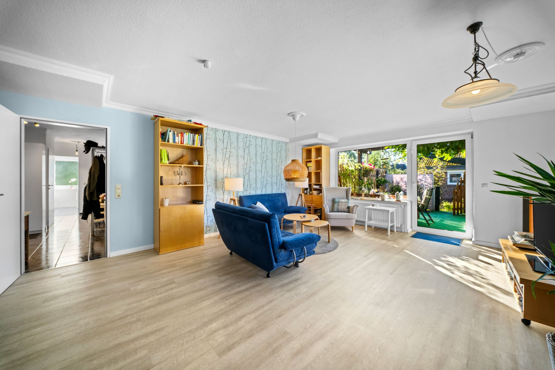 Helles Wohnzimmer mit Holzboden, blauer Couch, Bücherregalen und Zugang zur Terrasse mit Gartenblick.