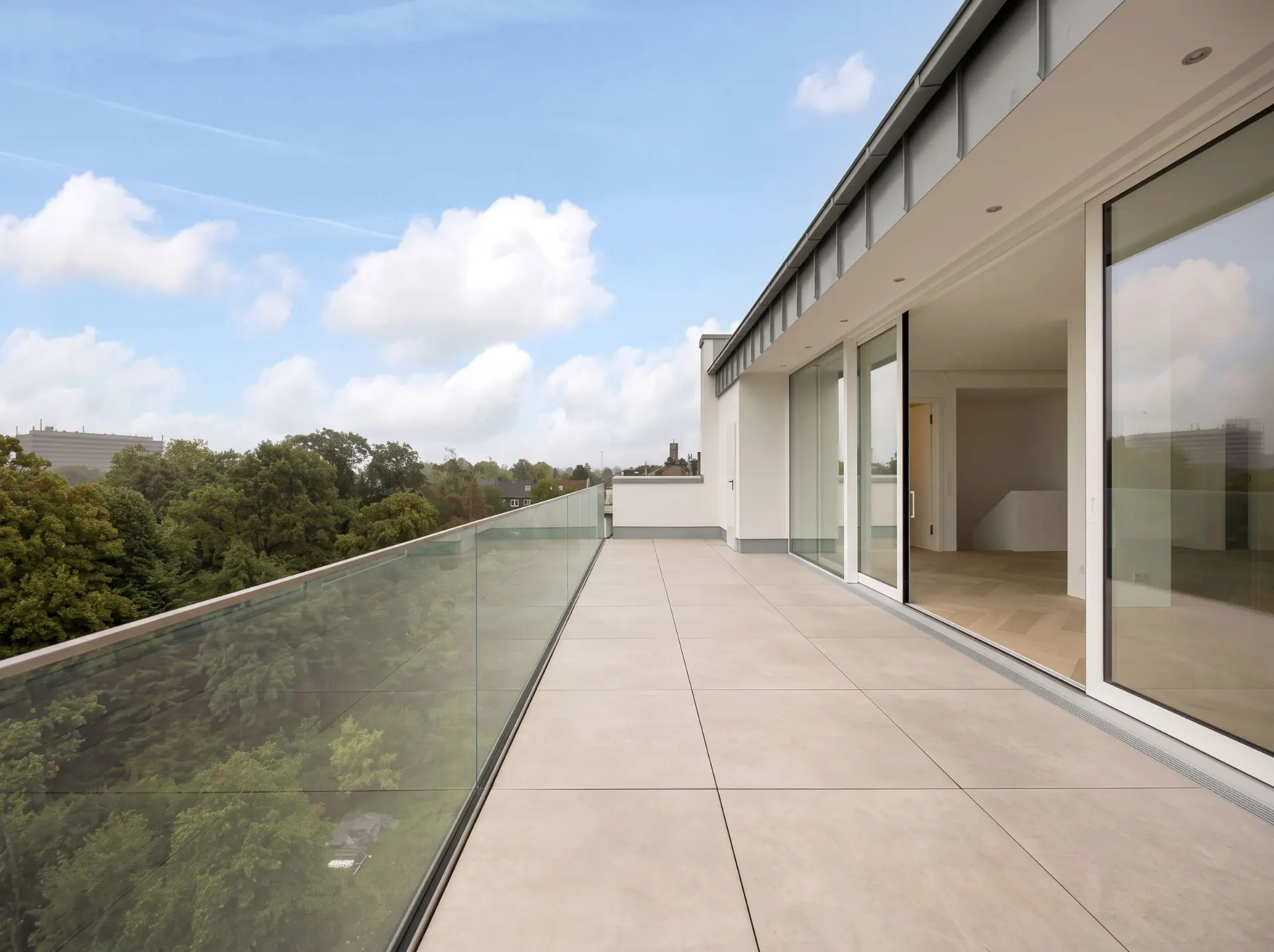 Große Terrasse mit Glasgeländer und Blick ins Grüne, angrenzend an Wohnräume mit großen Fenstern.