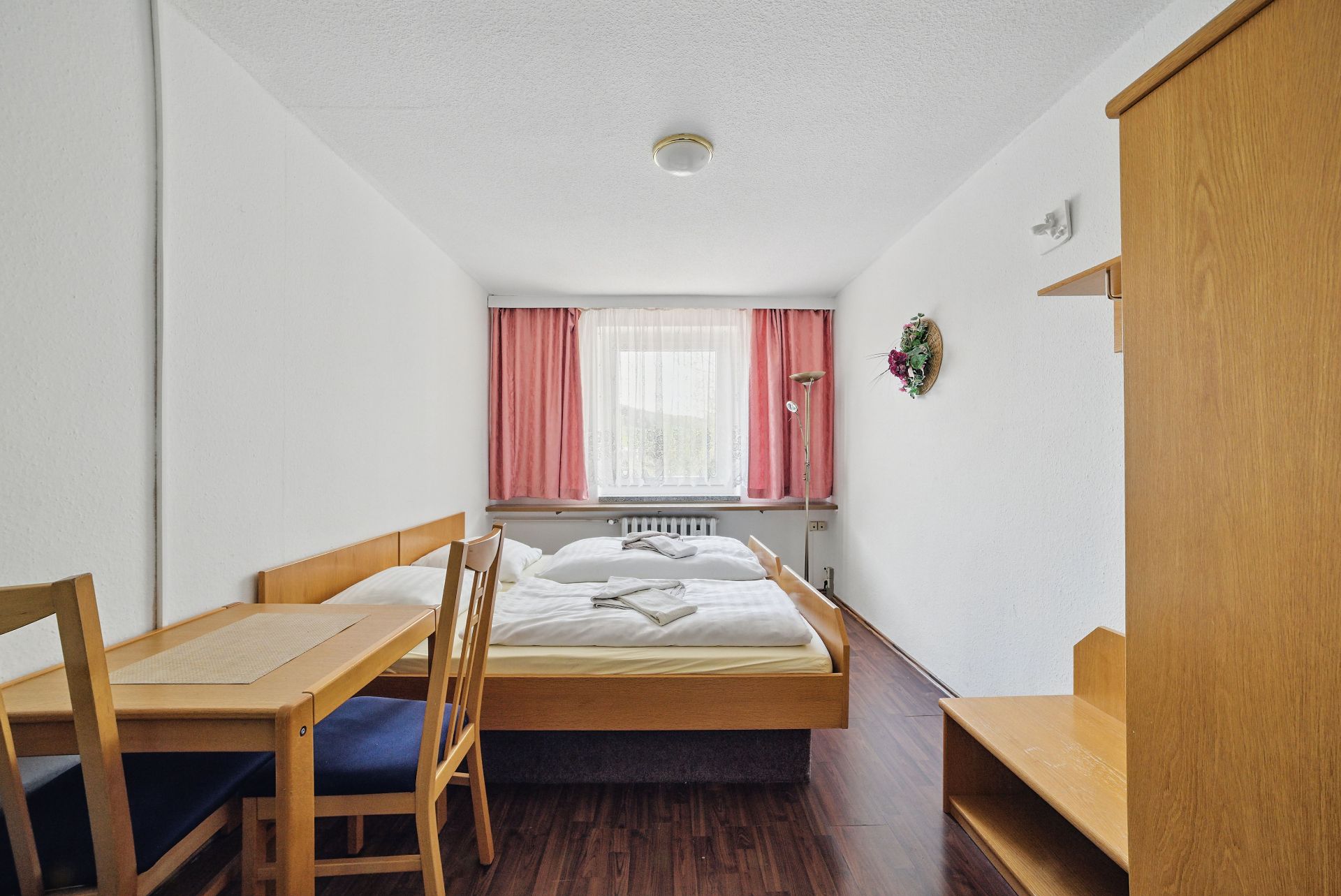 Schlafzimmer mit Doppelbett, Tisch, zwei Stühlen und Fenster mit rosa Vorhängen.