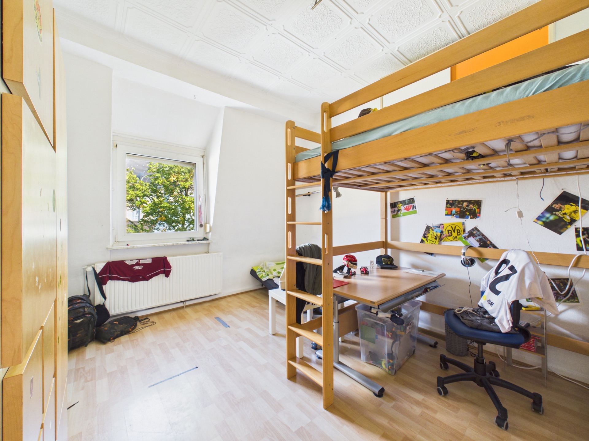 Helles Kinderzimmer mit Hochbett, Schreibtisch und Fenster, an den Wänden hängen Poster.