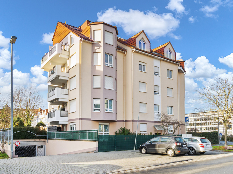 Mehrstöckiges Wohnhaus mit Balkonen, geparkten Autos und Zufahrt vor dem Gebäude.