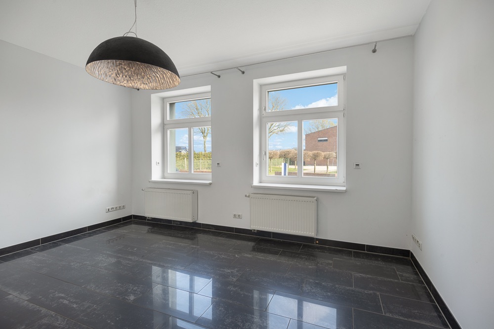 Leerer Raum mit zwei Fenstern, schwarzen Fliesen und einer großen Deckenlampe.