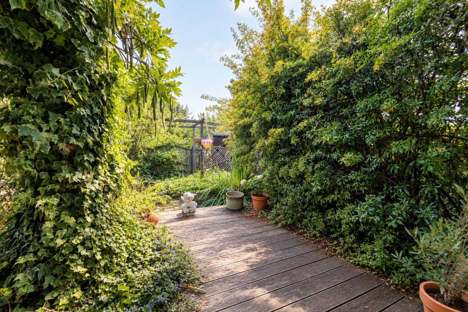 Garten mit Holzterrasse, vielen Pflanzen, Blumenkübeln und kleiner Gartenfigur.