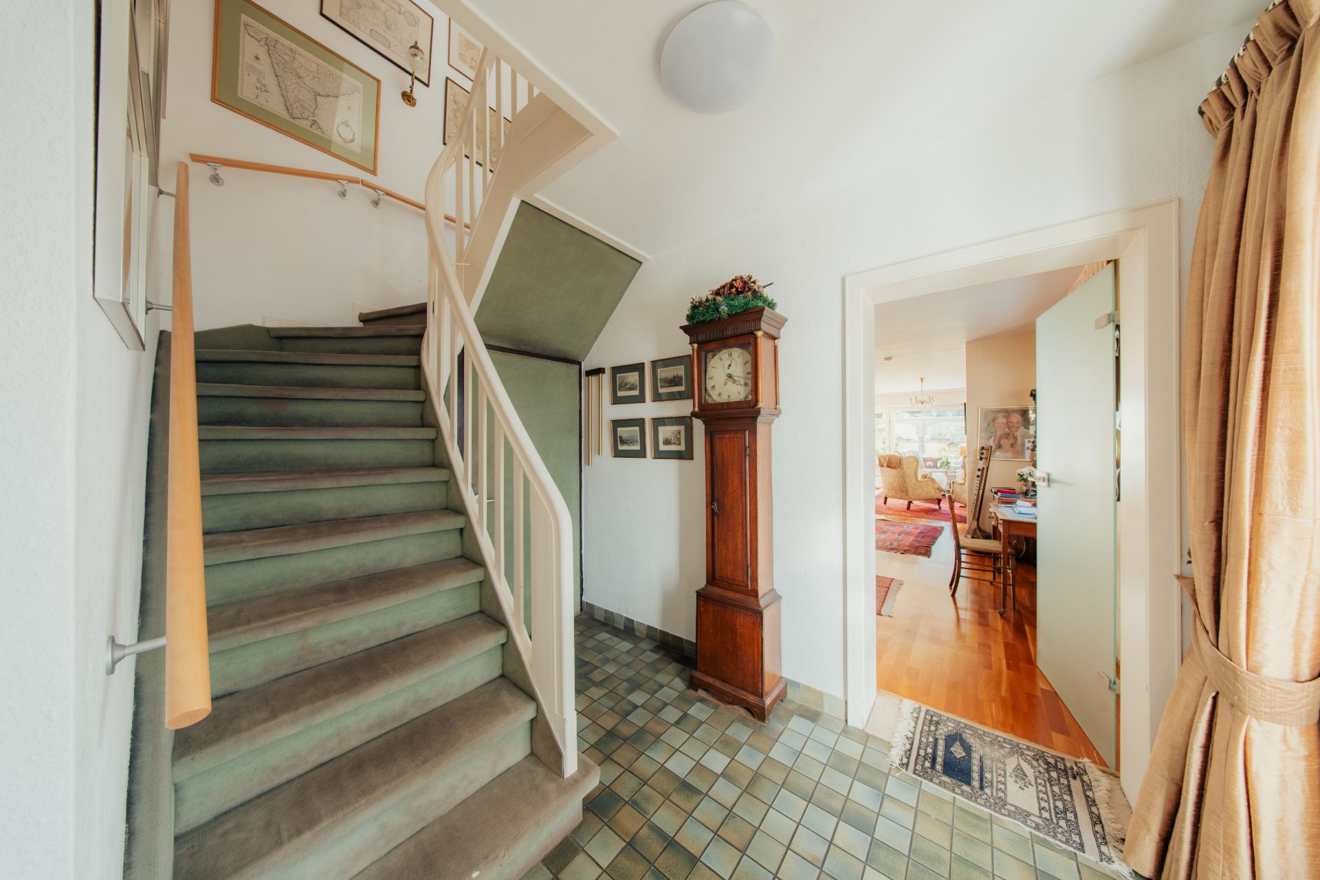 Flur mit Teppichtreppe, Wanduhr, Fliesenboden und Blick in ein Wohnzimmer.