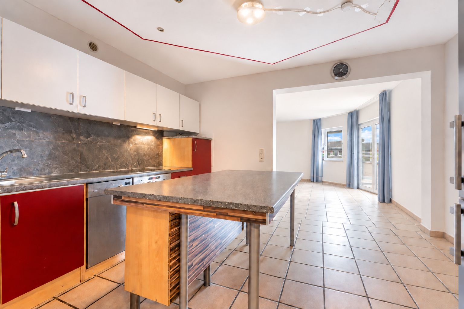 Helle Küche mit Kochinsel, roten und weißen Schränken, Fliesenboden und Blick ins Wohnzimmer.