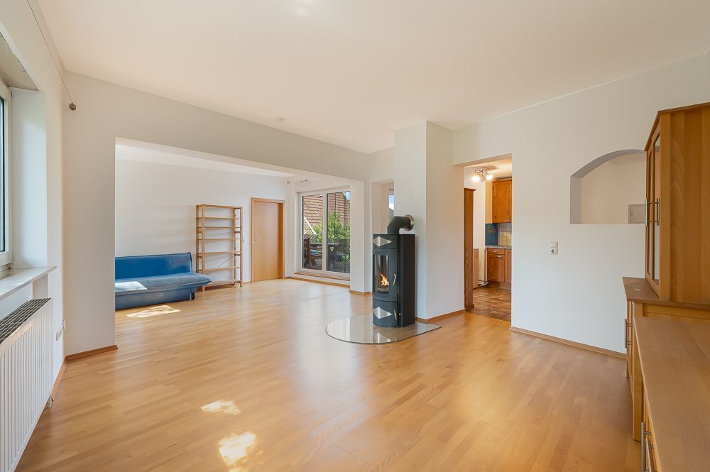 Helles Wohnzimmer mit Holzboden, Kaminofen, Sofa, Regal und Zugang zur Küche und Terrasse.