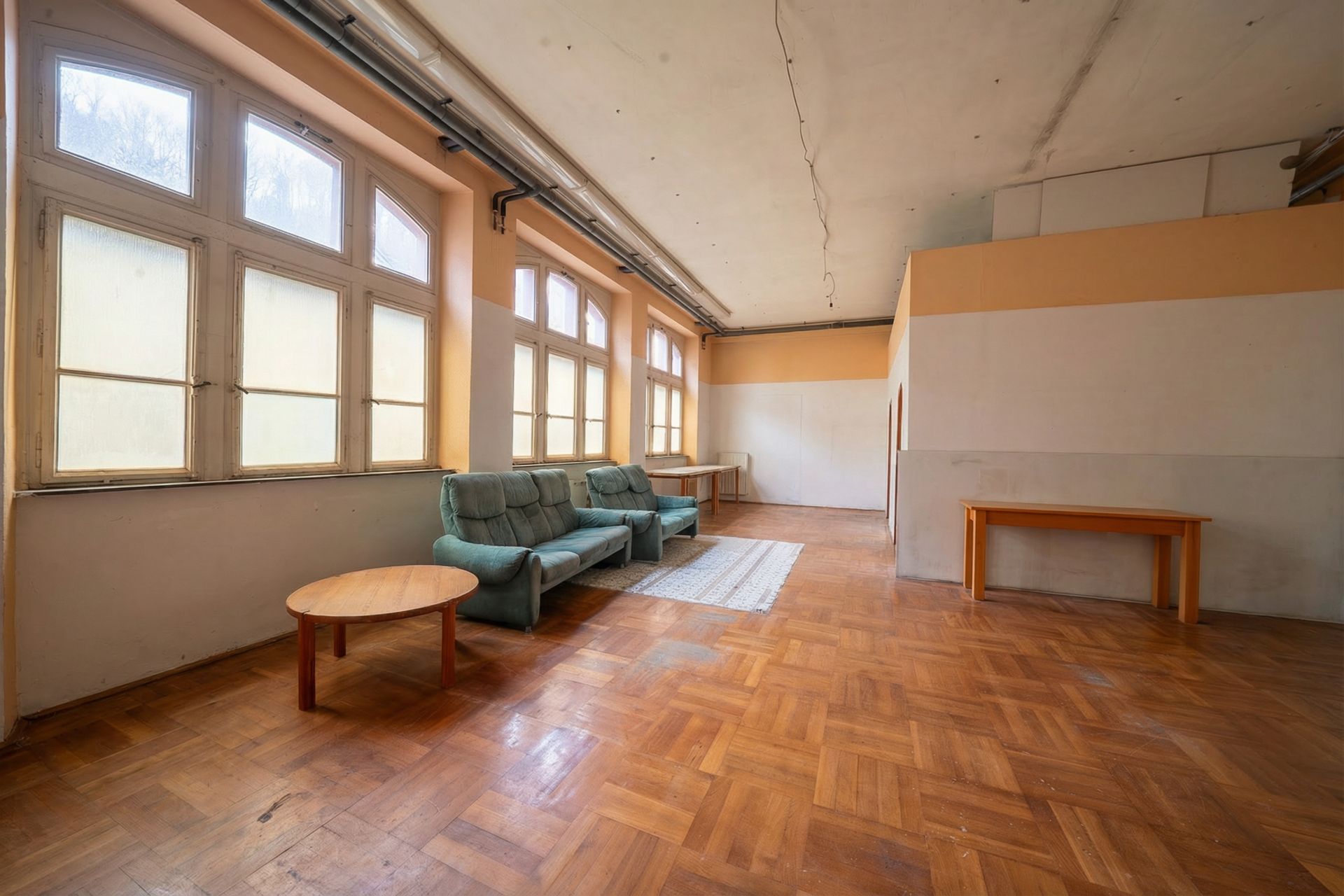 Großer Raum mit Parkettboden, Sofa, Couchtisch, Teppich und mehreren großen Fenstern.