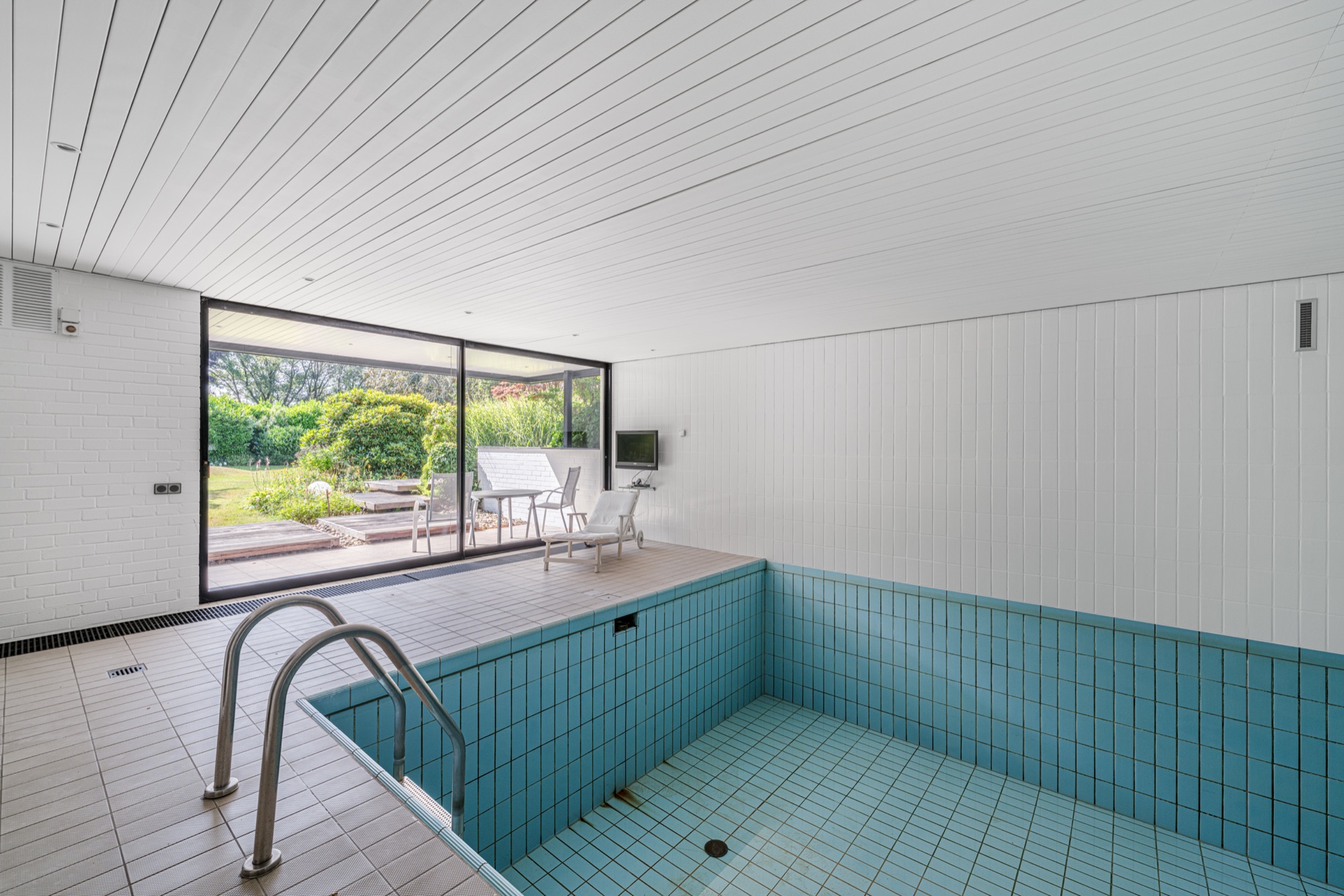 Leerer Innenpool mit blauen Fliesen, Sitzbereich und großen Fenstern zum Garten.