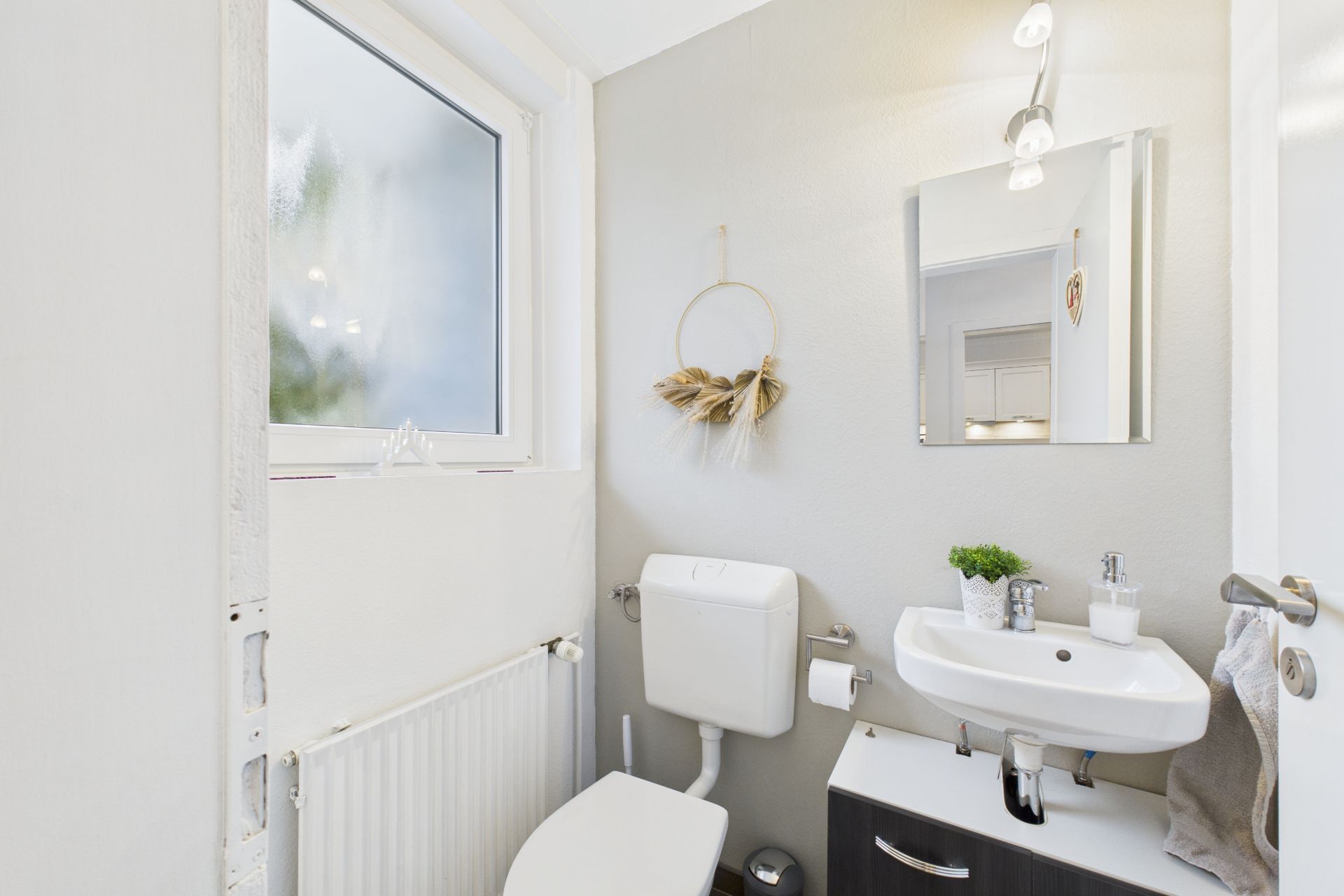 Kleines helles Badezimmer mit Fenster, Toilette, Waschbecken, Spiegel und Pflanzen.