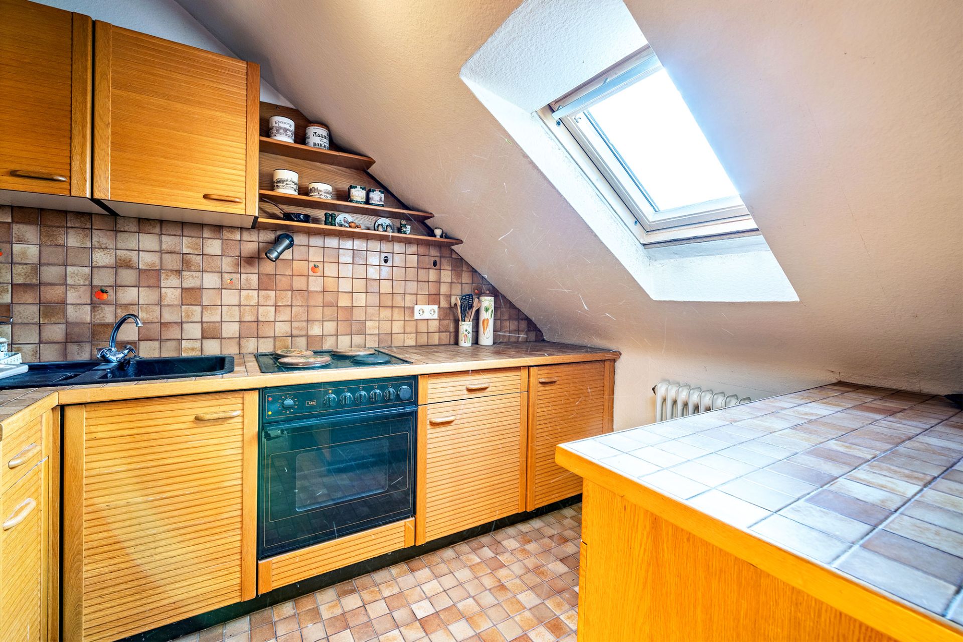 Küche mit Holzschränken, Schrägdachfenster und Fliesen auf Arbeitsfläche und Boden.