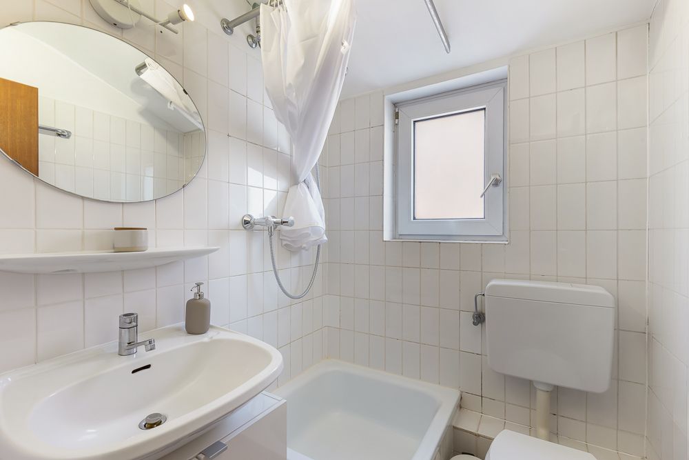 Kleines Badezimmer mit Waschbecken, Spiegel, Dusche, WC und Fenster.