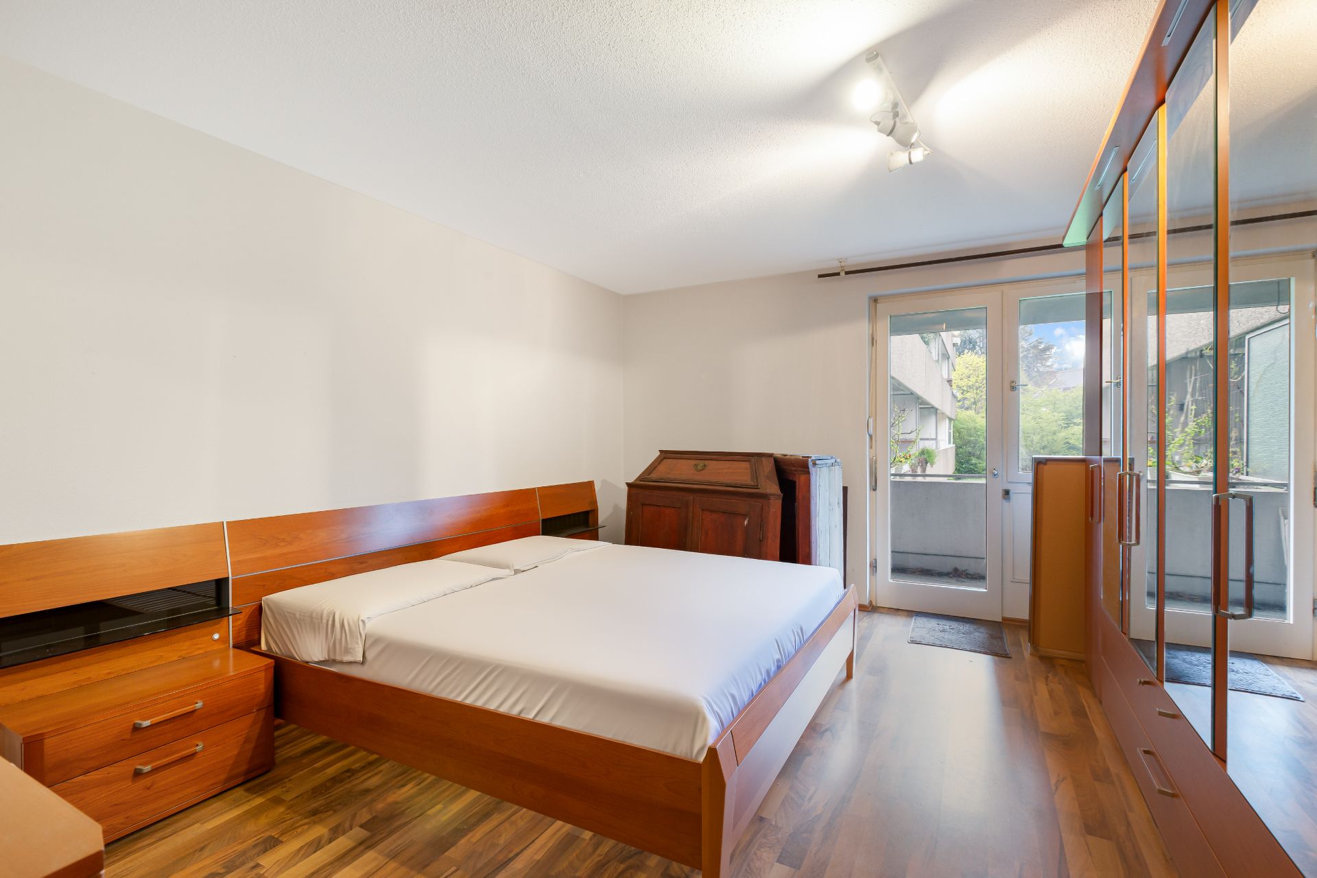 Schlafzimmer mit Holzbett, Schrank, Schreibtisch und Zugang zum Balkon.