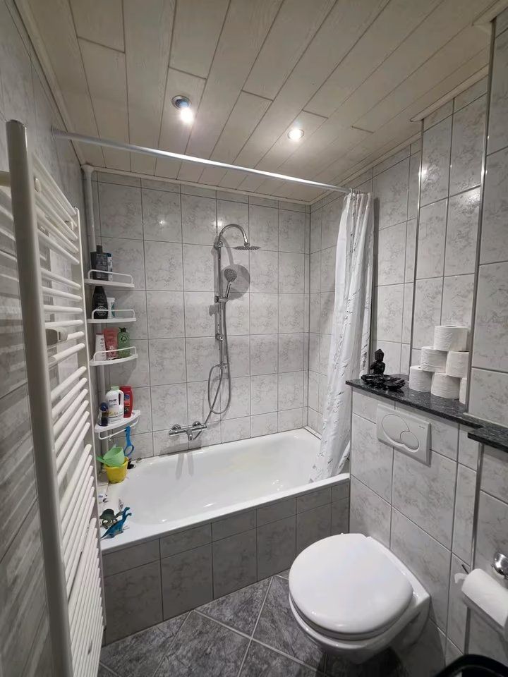 Kleines Badezimmer mit Badewanne, Dusche, WC, Regal und Fliesen an Wand und Boden.