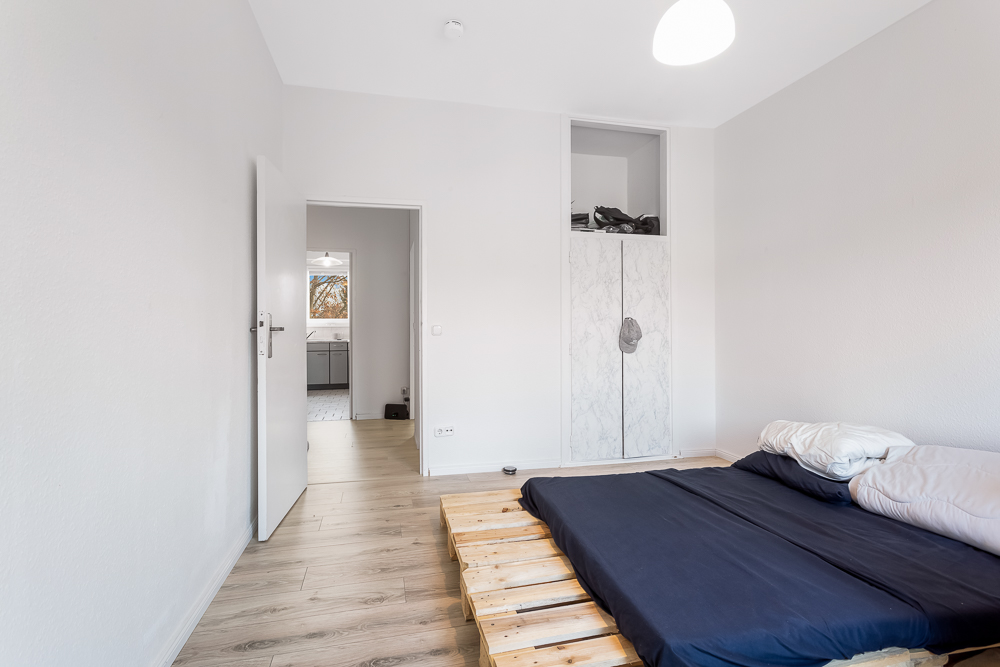 Schlafzimmer mit Palettenbett, blauer Bettwäsche und offenem Kleiderschrank, Blick zur Küche.