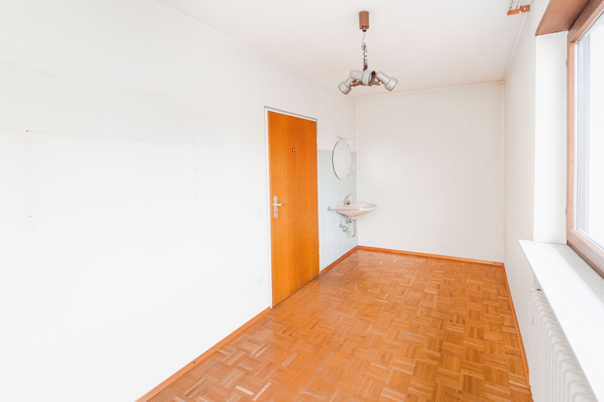 Leerer Raum mit Holzboden, Waschbecken, Spiegel, Tür und großem Fenster.