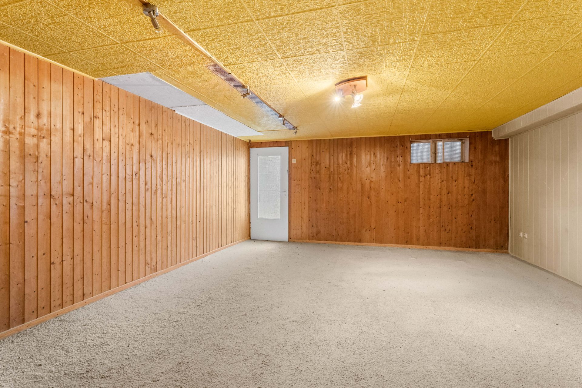 Kellerraum mit Holzvertäfelung, gelber Decke, Teppichboden, Tür und kleinem Fenster.