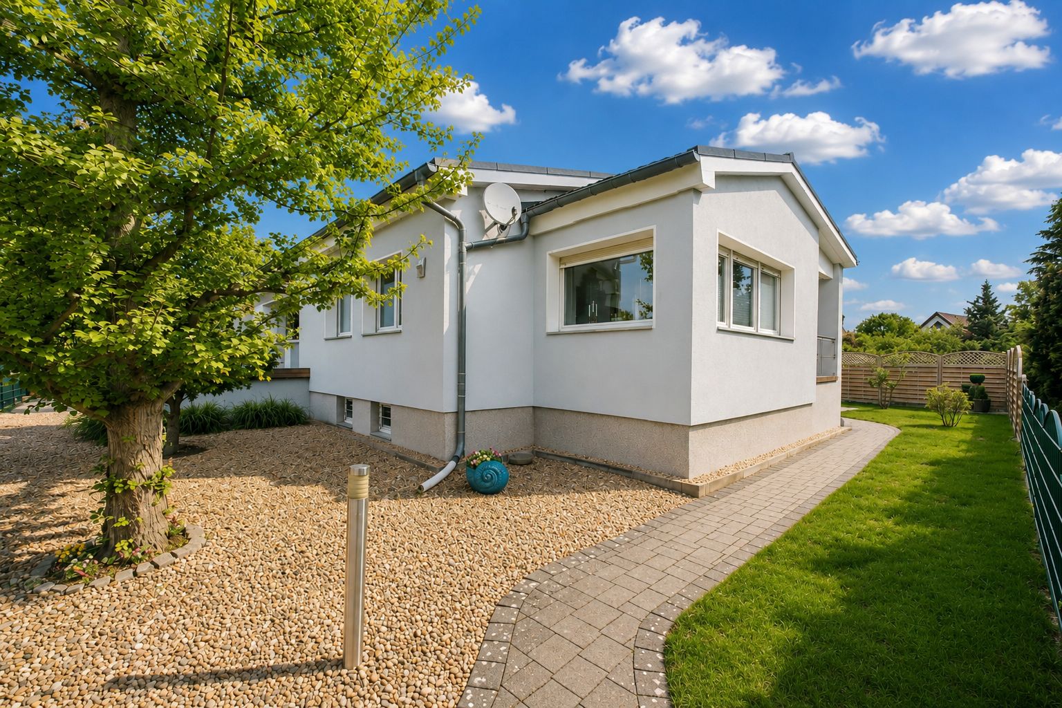 Einfamilienhaus mit weißen Wänden, Garten, Kiesweg und Baum bei sonnigem Wetter.