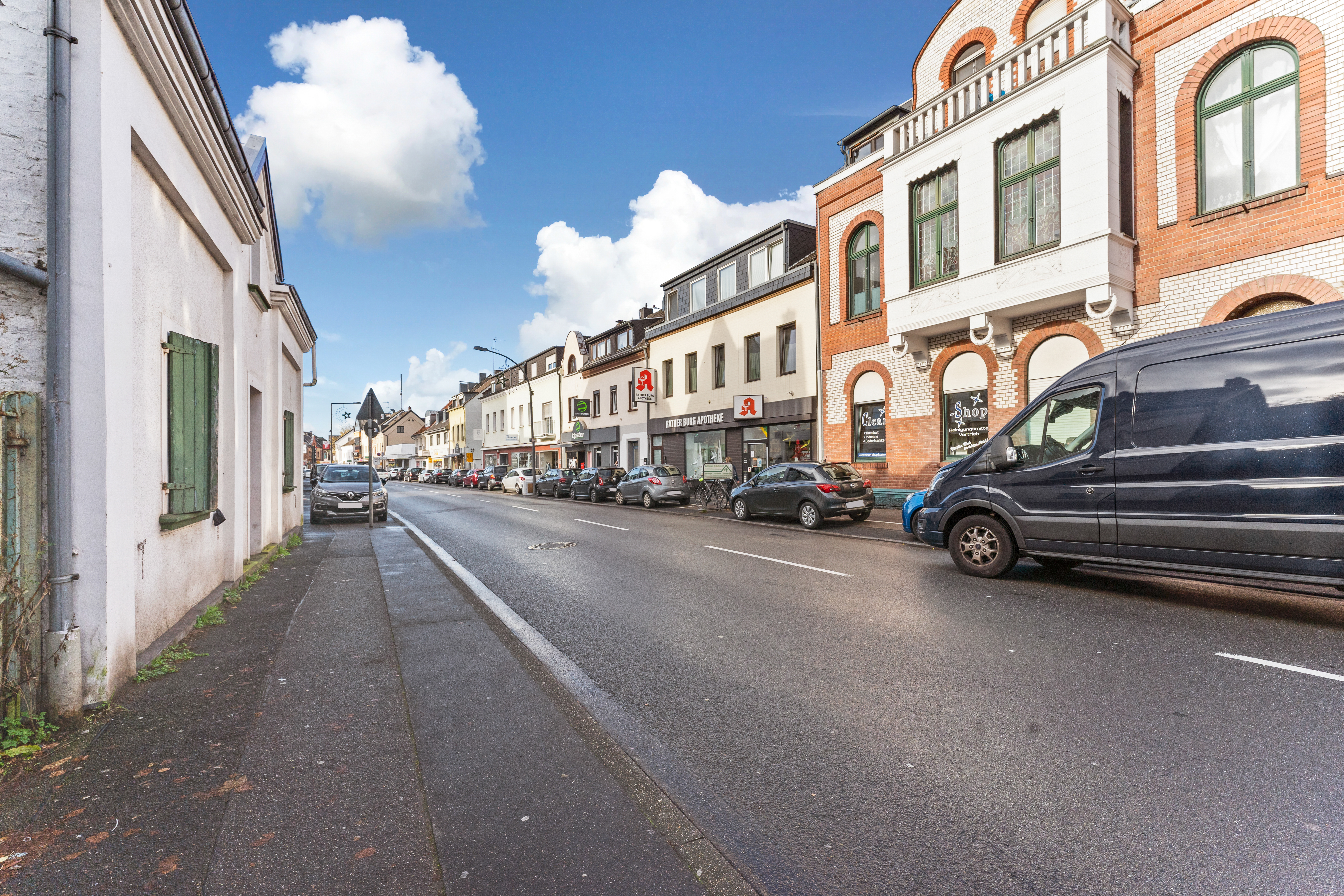 Straßenszene mit Mehrfamilienhäusern, parkenden Autos und Gehweg bei Tageslicht.