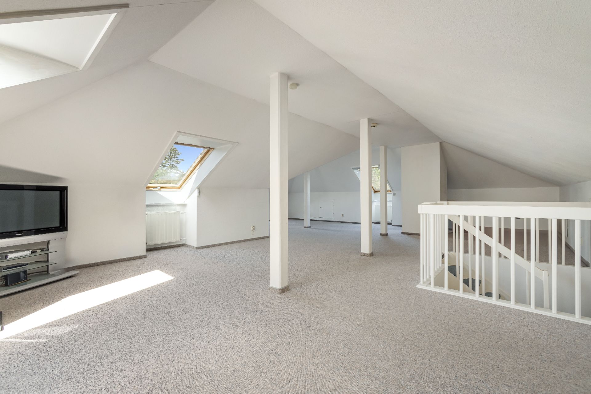 Großer, heller Dachbodenraum mit Teppichboden, TV, Dachfenstern und Treppe.