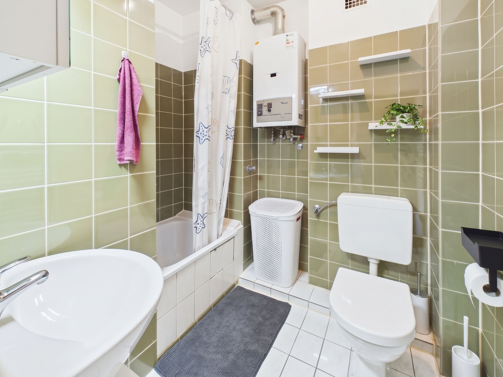 Helles Badezimmer mit grünen Fliesen, Badewanne, Waschbecken, Toilette und Regal mit Pflanze.