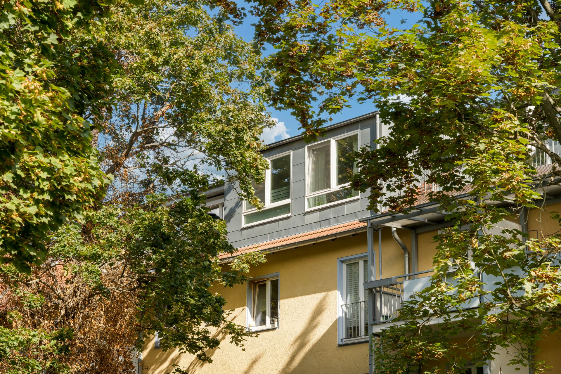 Wohnhaus mit gelber Fassade und Balkon, teilweise von grünen Bäumen verdeckt.