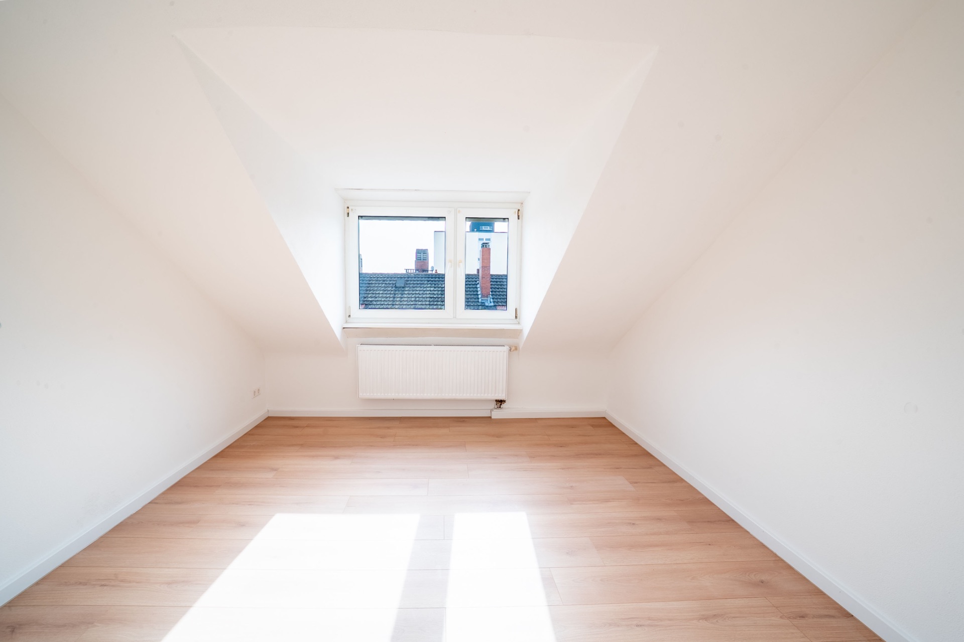 Heller Dachraum mit Holzfußboden, weiß gestrichenen Wänden und einem Fenster mit Blick auf Dächer.