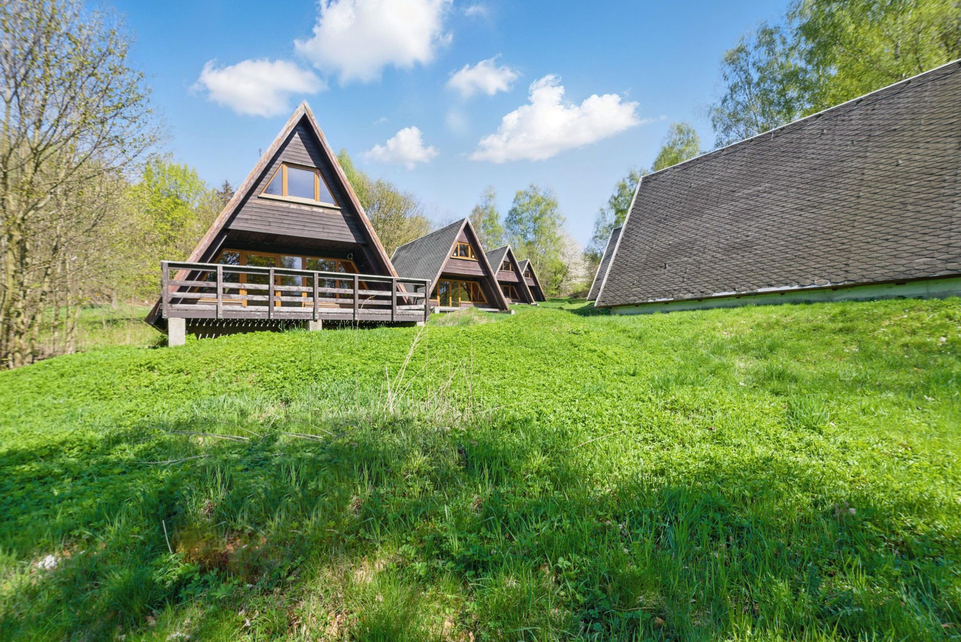 Dreieckige Holzhäuser mit Terrasse stehen auf einer grünen Wiese, umgeben von Bäumen.