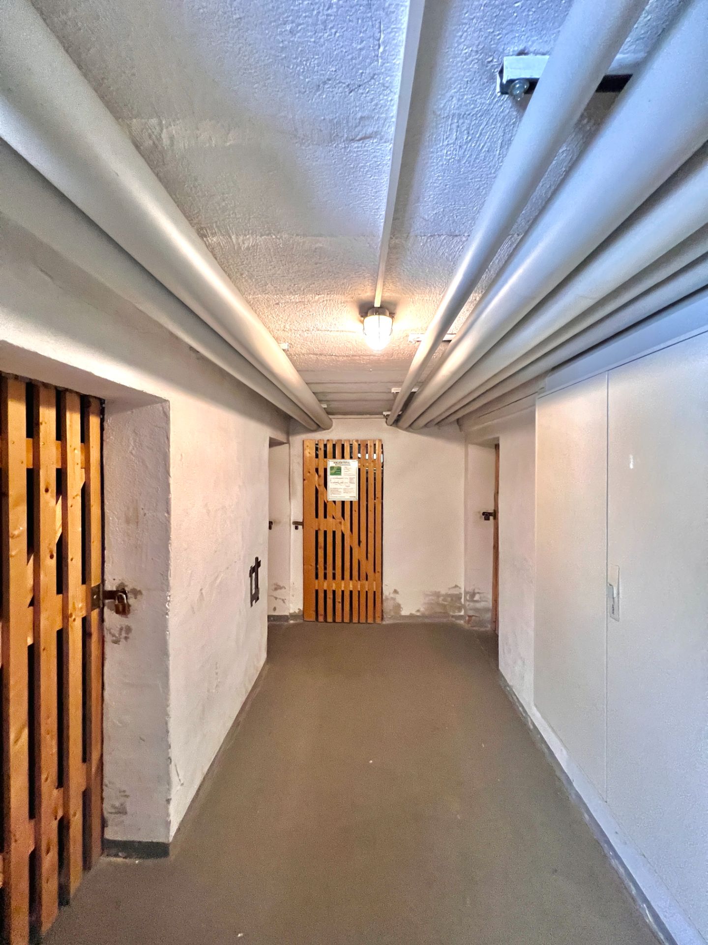 Heller Kellerflur mit Holzgittertüren, Rohren an der Decke und einer Lampe.