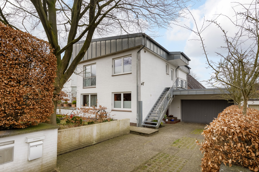 Zweistöckiges Wohnhaus mit weißer Fassade, Außentreppe und Garage, umgeben von Büschen und Bäumen.