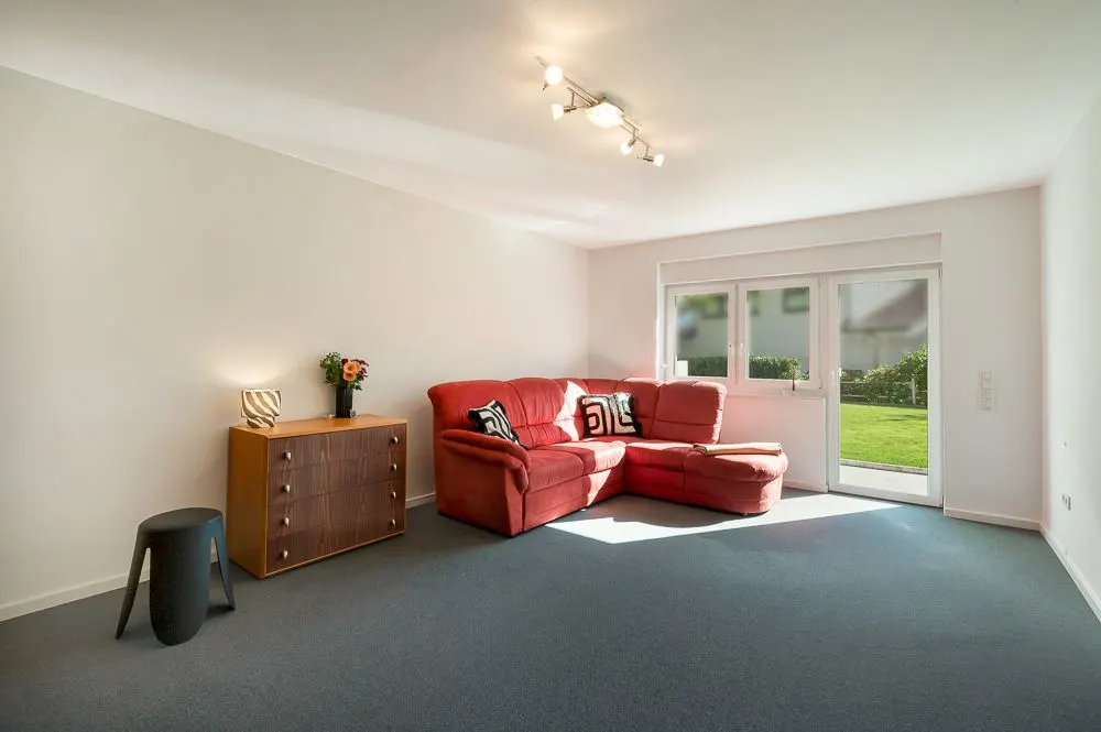 Helles Wohnzimmer mit rotem Ecksofa, Kommode, Hocker und Blick auf den Garten durch große Fenster.
