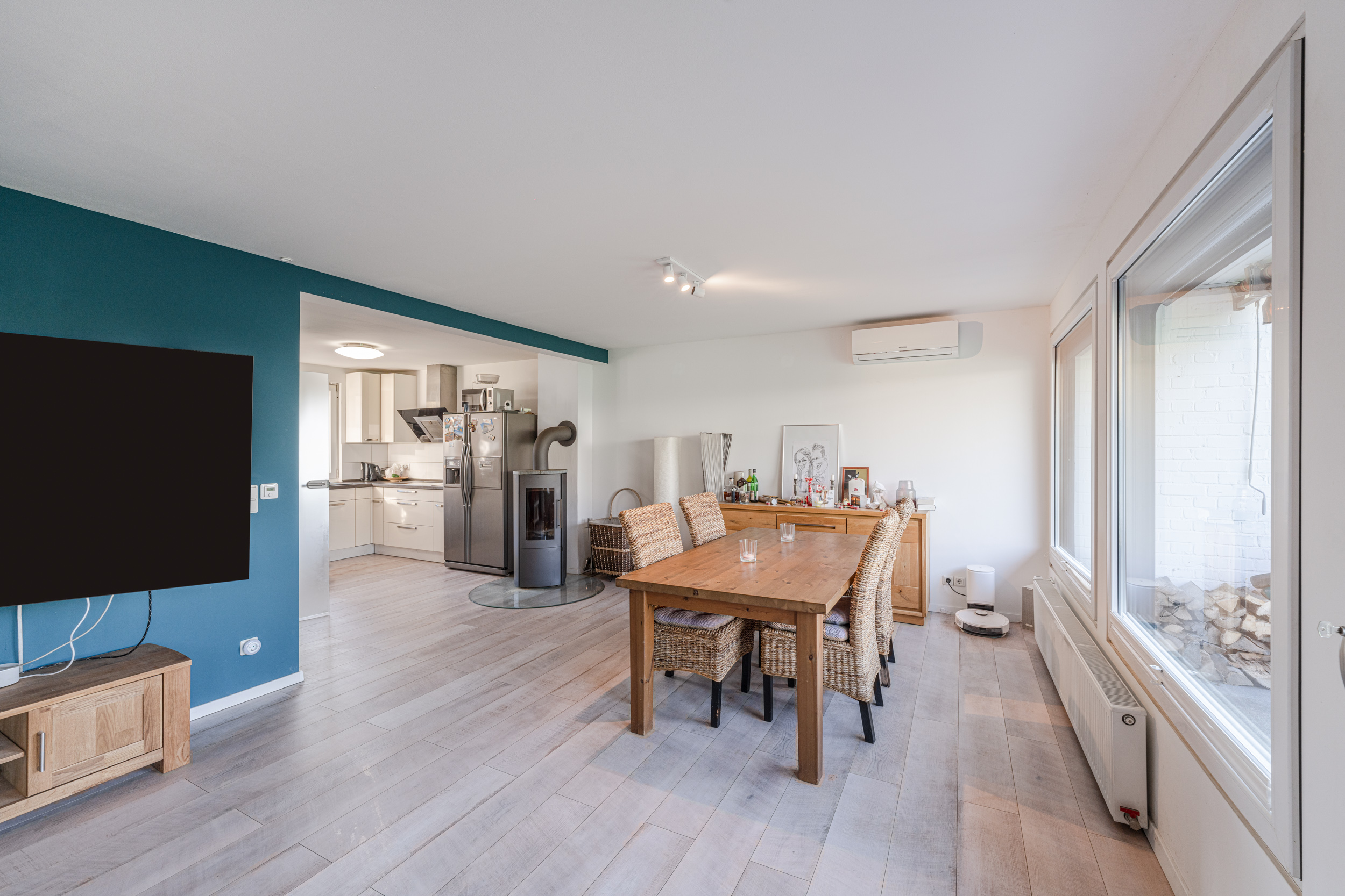 Helles Wohnzimmer mit Esstisch, Stühlen, Fernseher, offener Küche und großen Fenstern.