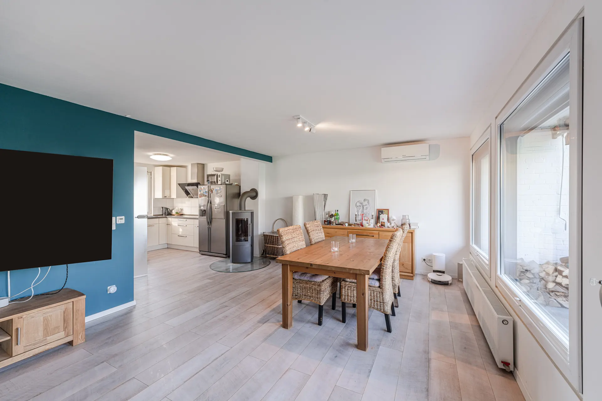 Helles Wohnzimmer mit Esstisch, Stühlen, Fernseher, offener Küche und großen Fenstern.