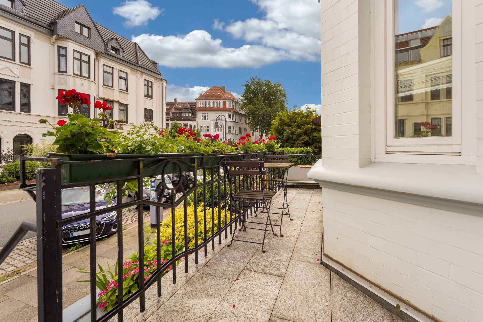 Möblierte Terrasse mit Blumen am Geländer, Blick auf Wohnhäuser und eine ruhige Straße.