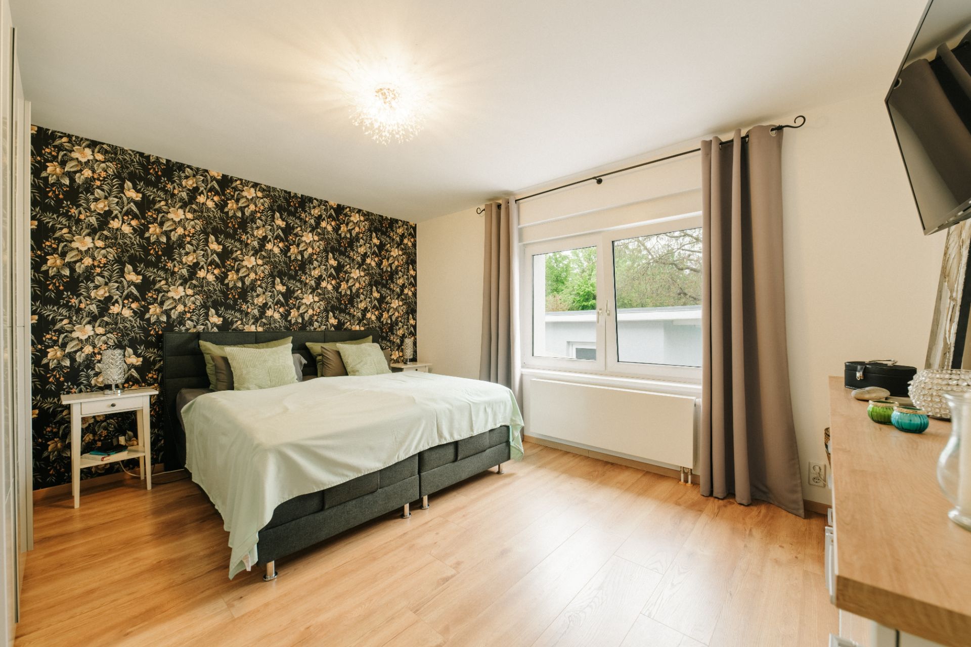 Helles Schlafzimmer mit Doppelbett, gemusterter Wand, Fenster und Holzboden.